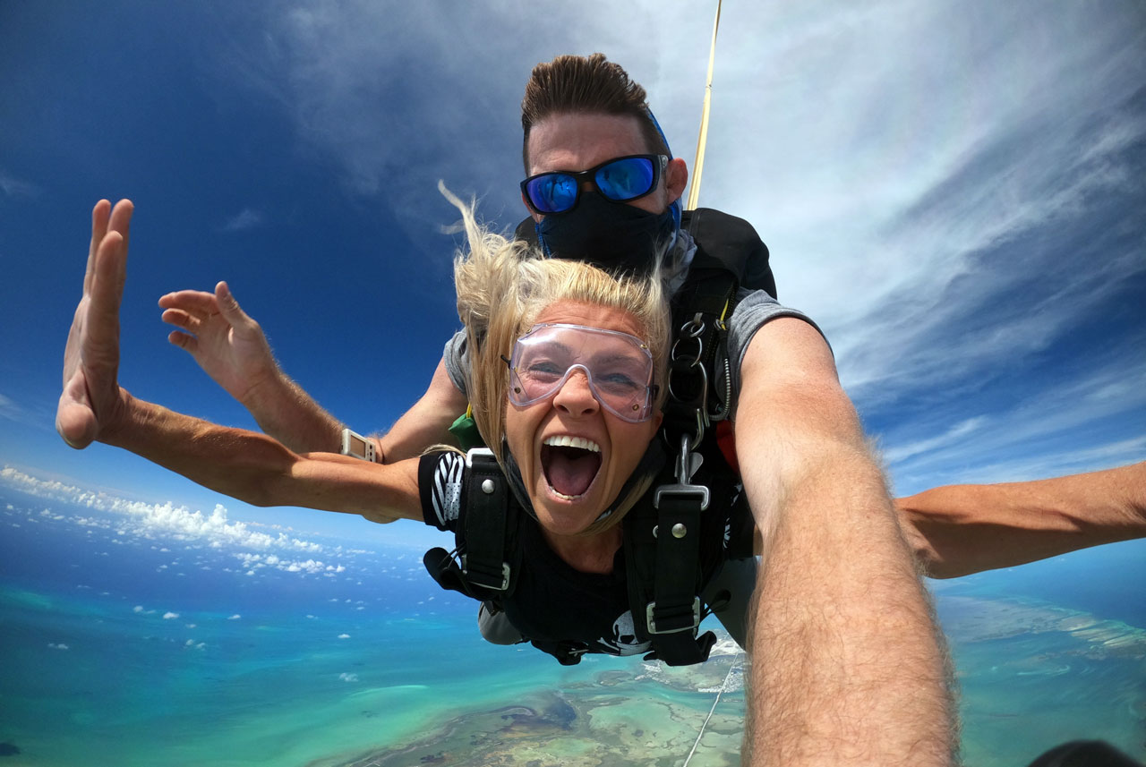 Female enjoying tandem skydiving outstretches her arm and smiles with glee during free fall