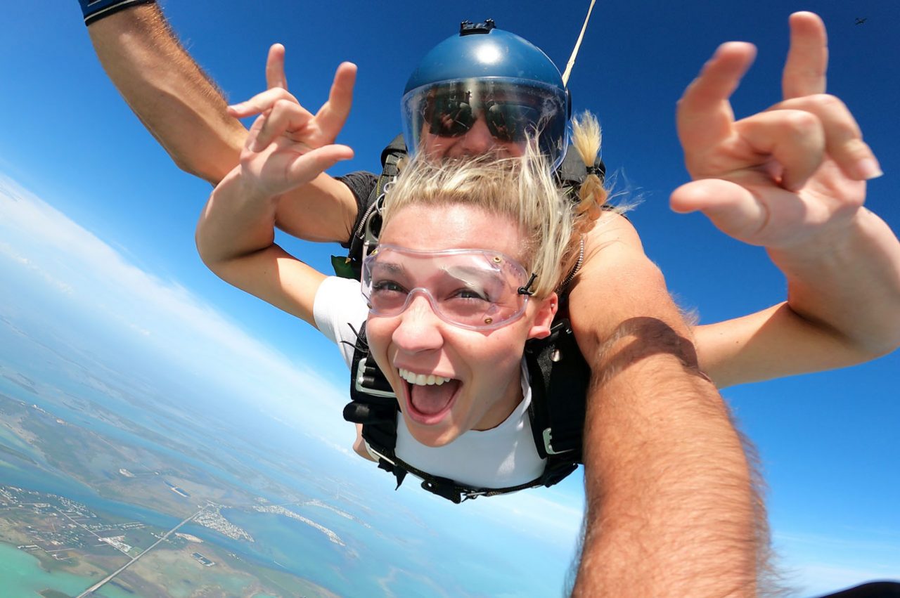 Young female wearing white shirt and clear goggles smiles during freefall at Skydive Key West