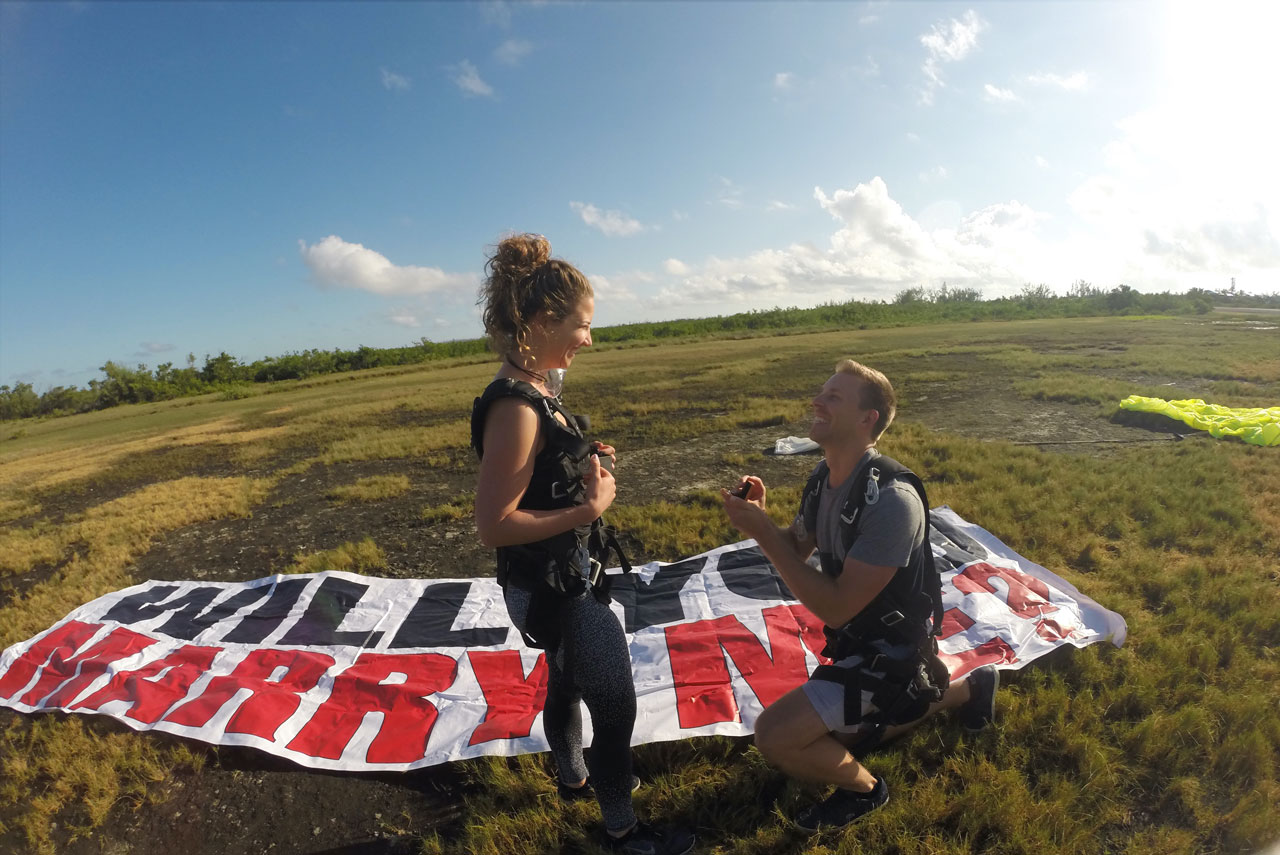 Young male gets down on one knee next to a "will you marry me" banner to purpose after Florida Keys Skydiving