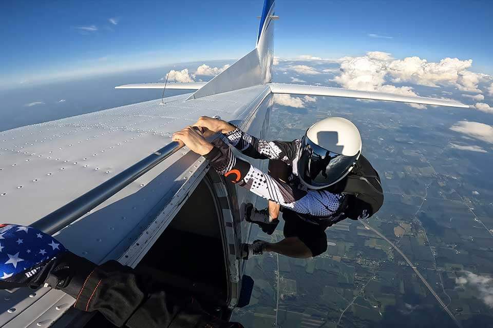 Skydiver in a white helmet perches on the side of an airplane prior to exit. 