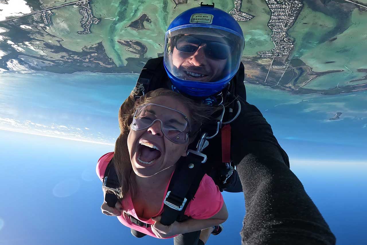 Female tandem skydiver in a pink shirt smiles big with her mouth open.