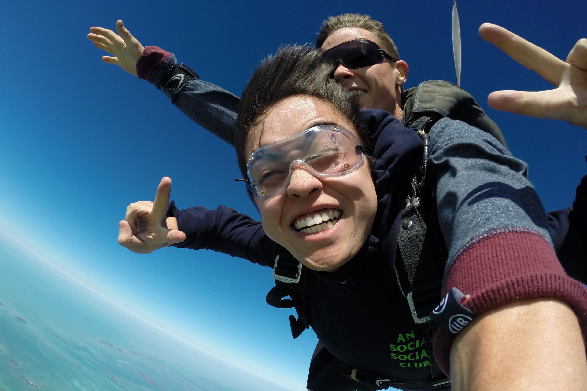 Smiling tandem skydiving student holds up two fingers on each hand during freefall to show peace signs.