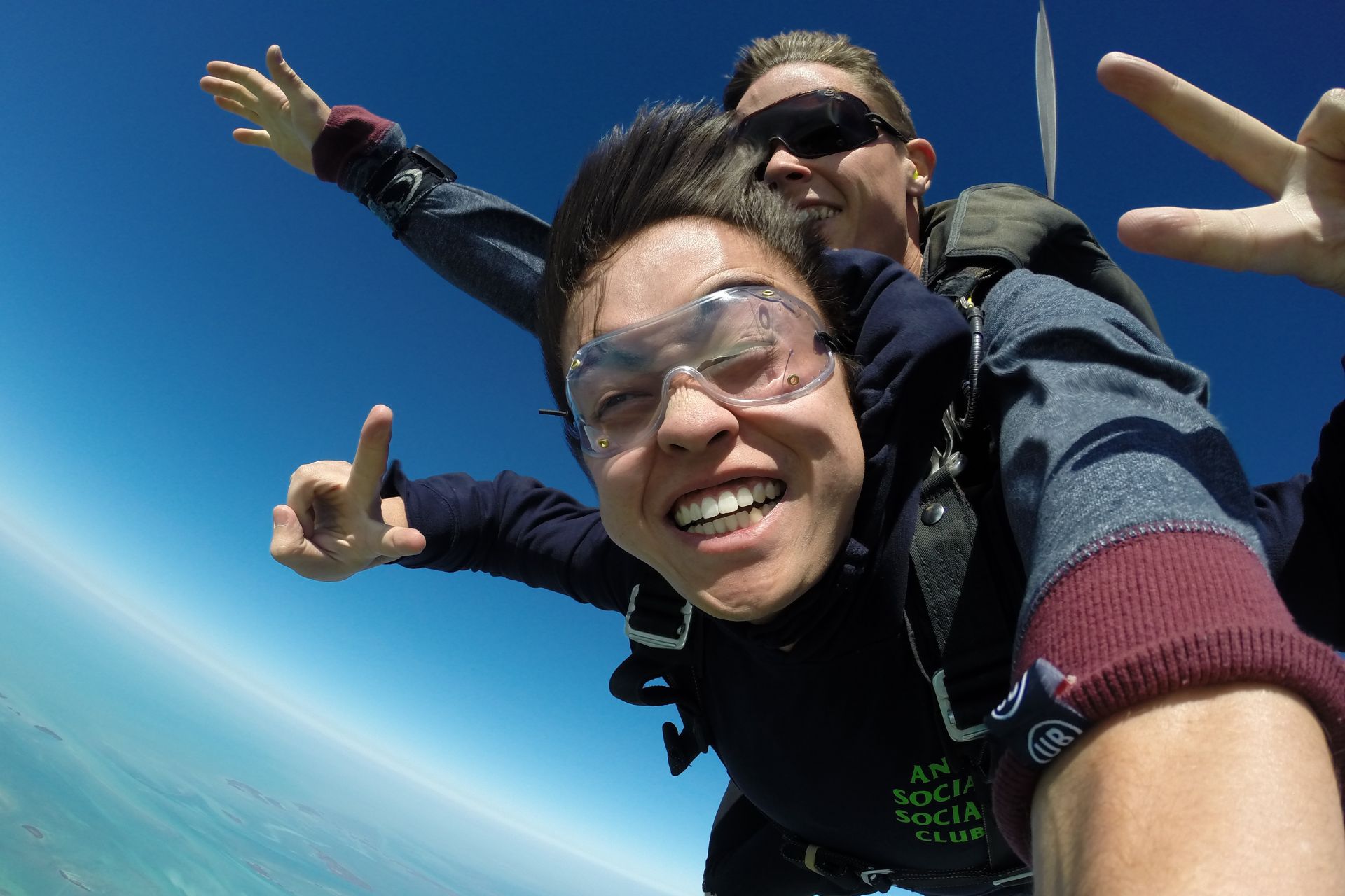 Smiling tandem skydiving student holds up two fingers on each hand during freefall to show peace signs. 